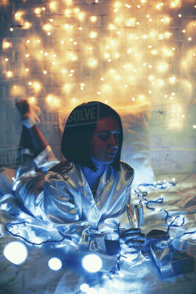 Thoughtful woman in pajamas with glass of champagne and gift lying on