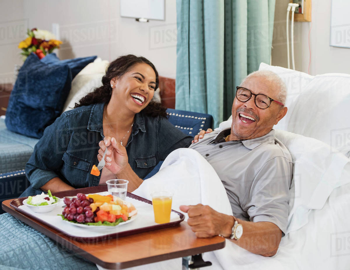 Laughing woman sitting by senior man eating fruit in bed - Stock Photo