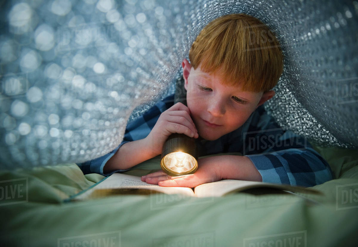 Boy (89) reading book under bed covers Stock Photo Dissolve