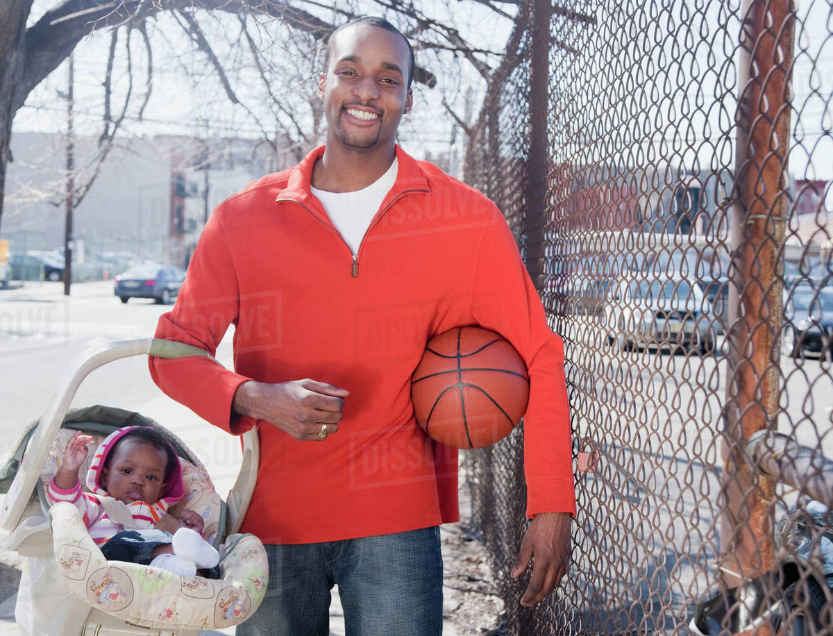 African man carrying baby in carrier and holding basketball Stock