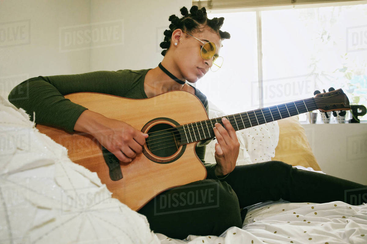 Mixed Race woman sitting on bed playing guitar Stock Photo Dissolve