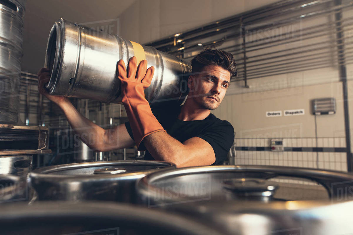 Shot of brewer carrying keg at brewery factory. Young man with metal