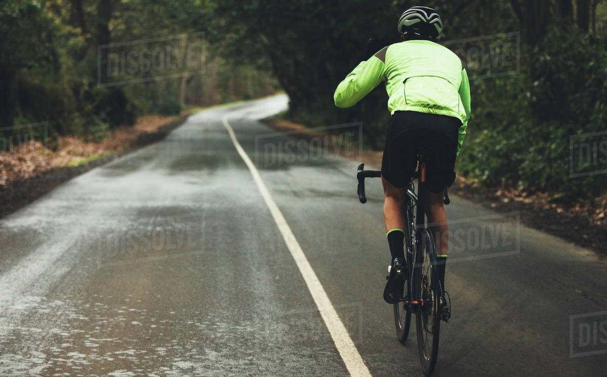 Rear view of male athlete cycling on country road on rainy day. Professional cyclist riding a bike on empty highway through forest. Royalty-free stock photo