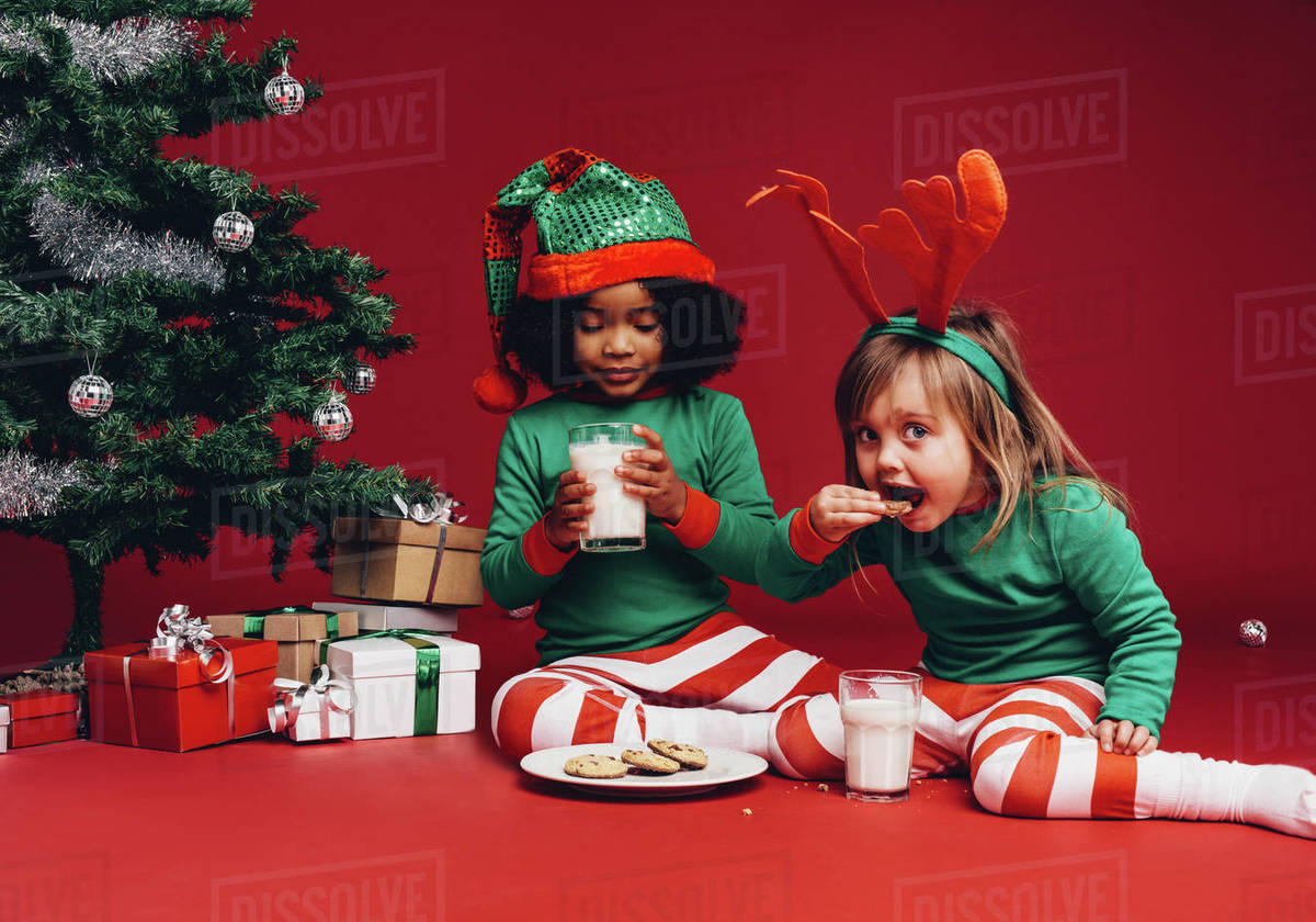 Two kids sitting beside a christmas tree eating cookies with milk