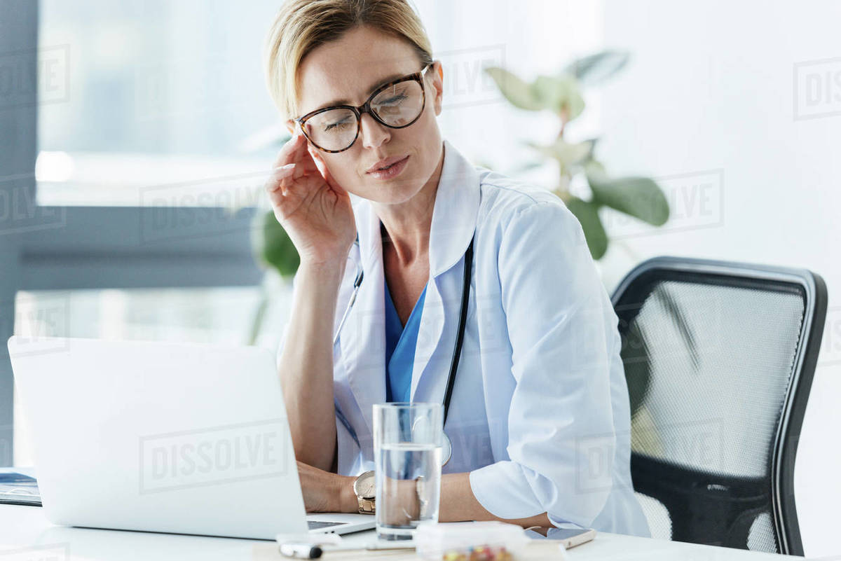 Exhausted female doctor in eyeglasses having headache at table in office Stock Photo Dissolve