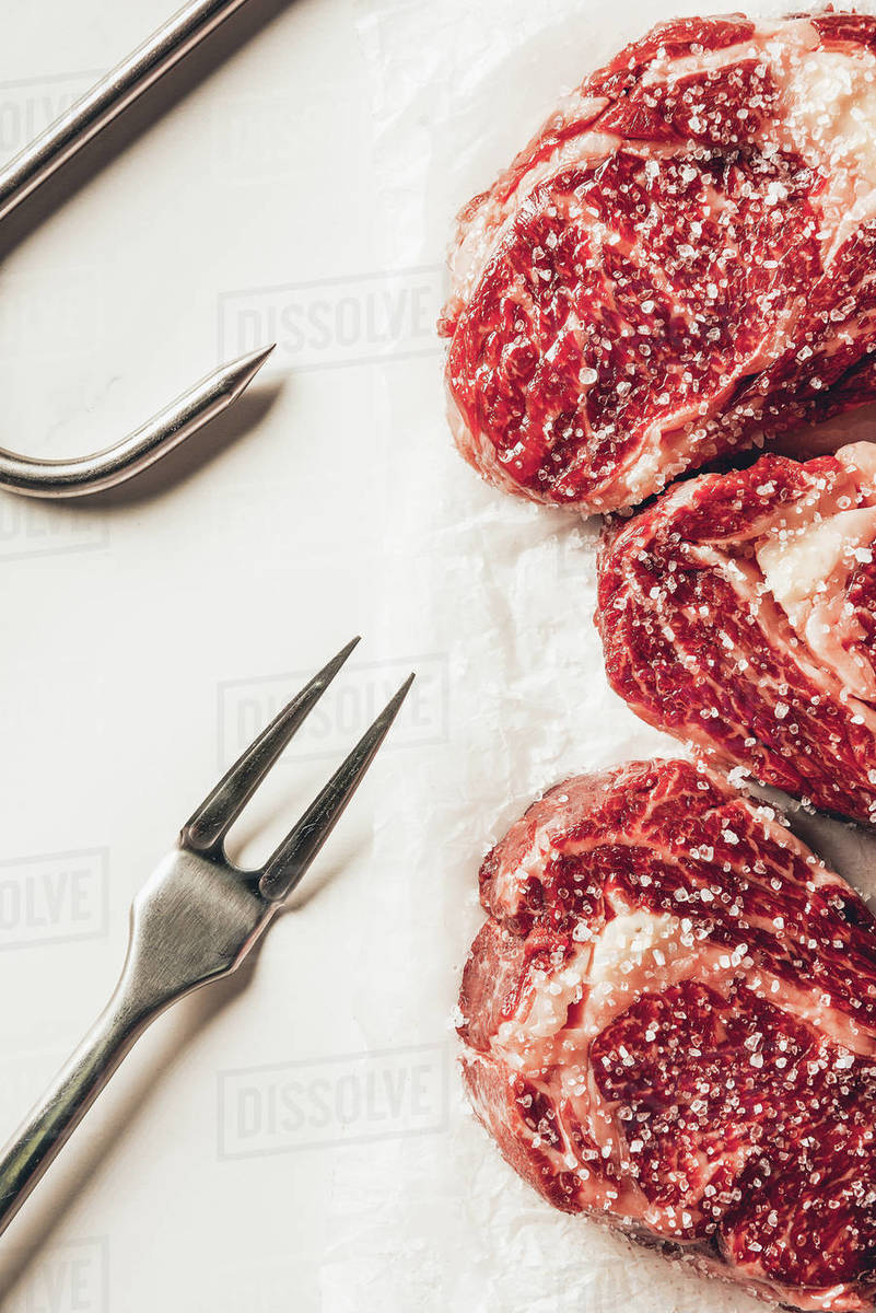 Top View Of Three Pieces Of Raw Meat Steaks With Salt Meat Fork And Metal Hook On White Surface 