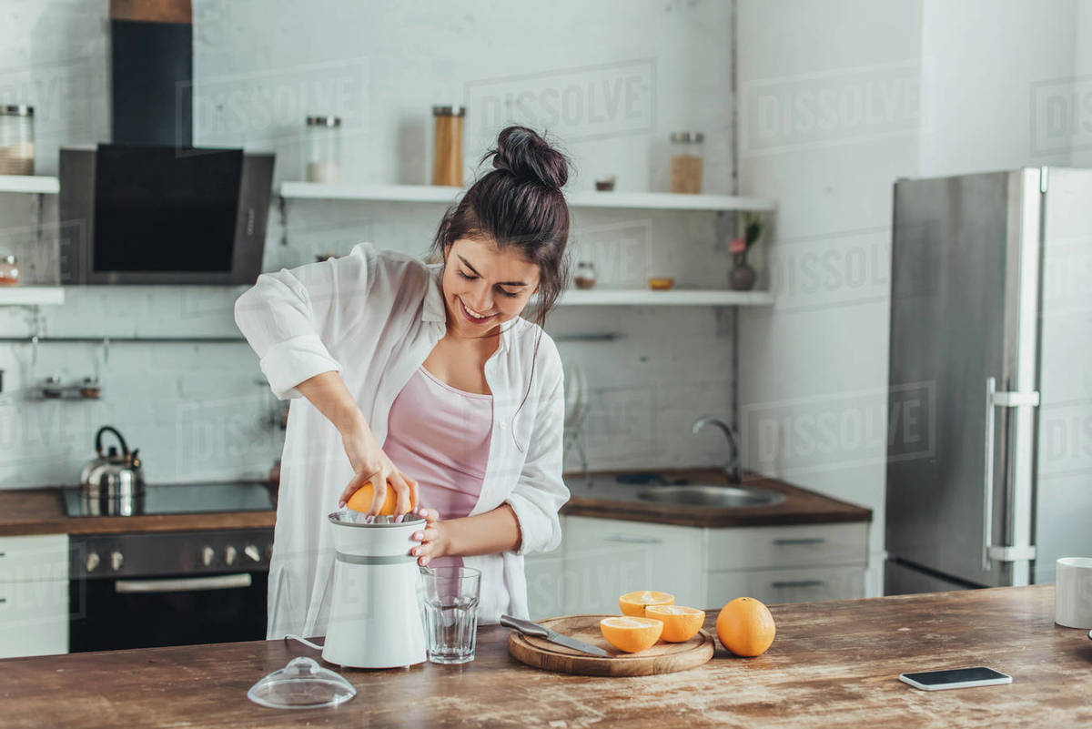 Smiling girl making fresh orange juice with juicer at wooden table in