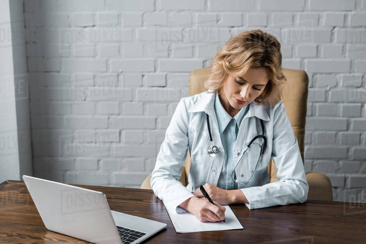 Confident female doctor writing document at workplace in office Stock Photo Dissolve
