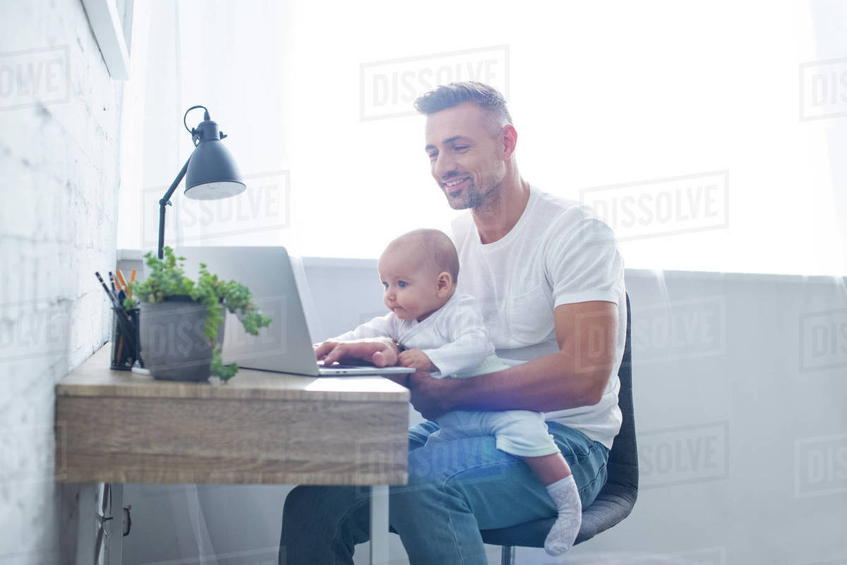 Smiling father sitting on chair, holding baby and using laptop at home