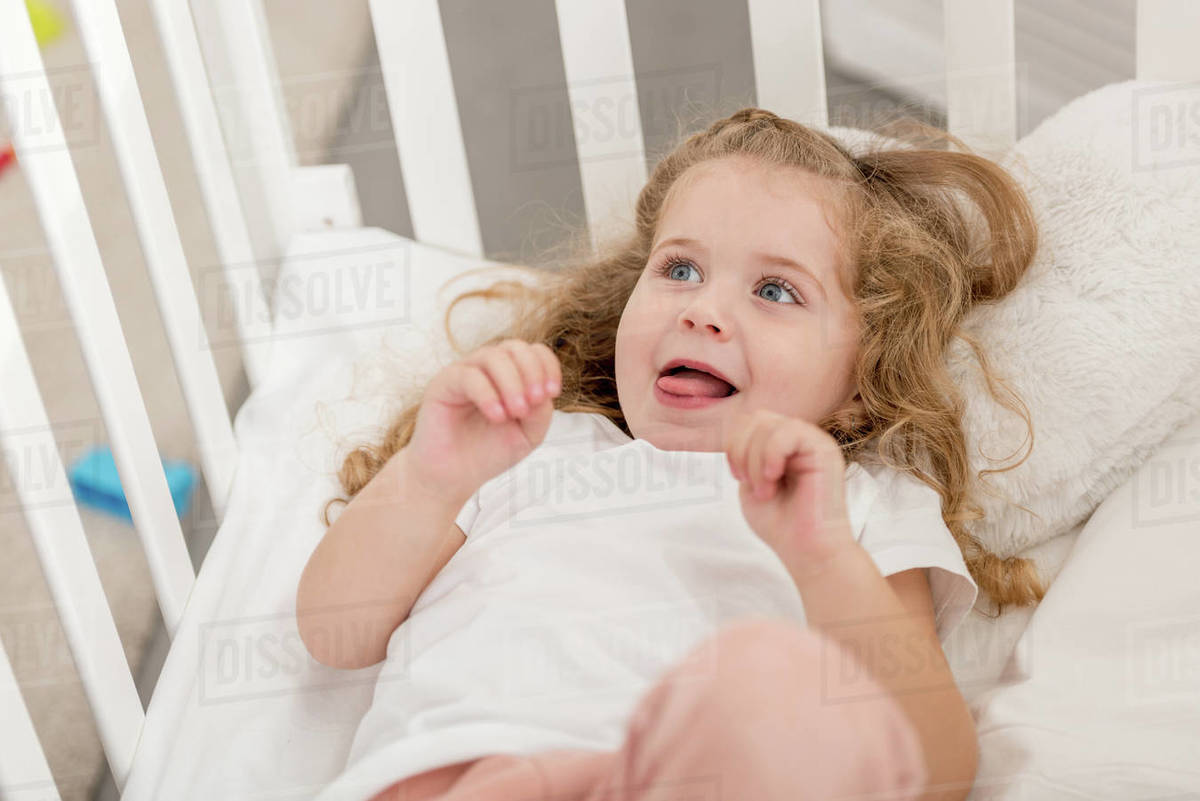 Adorable kid sticking tongue out and lying in crib in children room