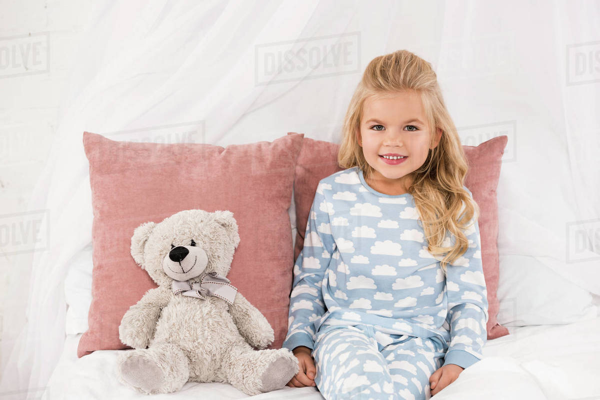 Cute Smiling Child Sitting On Bed With Teddy Bear And Pillows