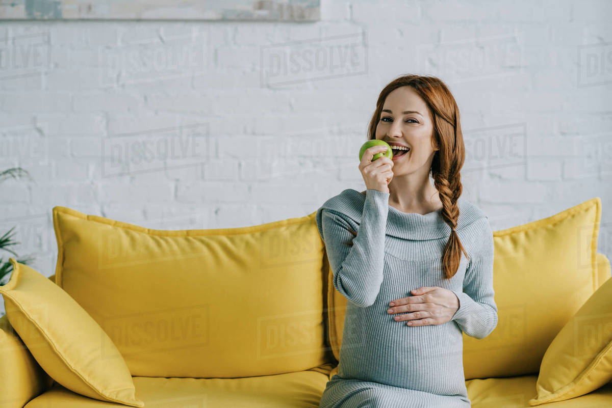 Happy attractive pregnant woman eating apple in living room Stock