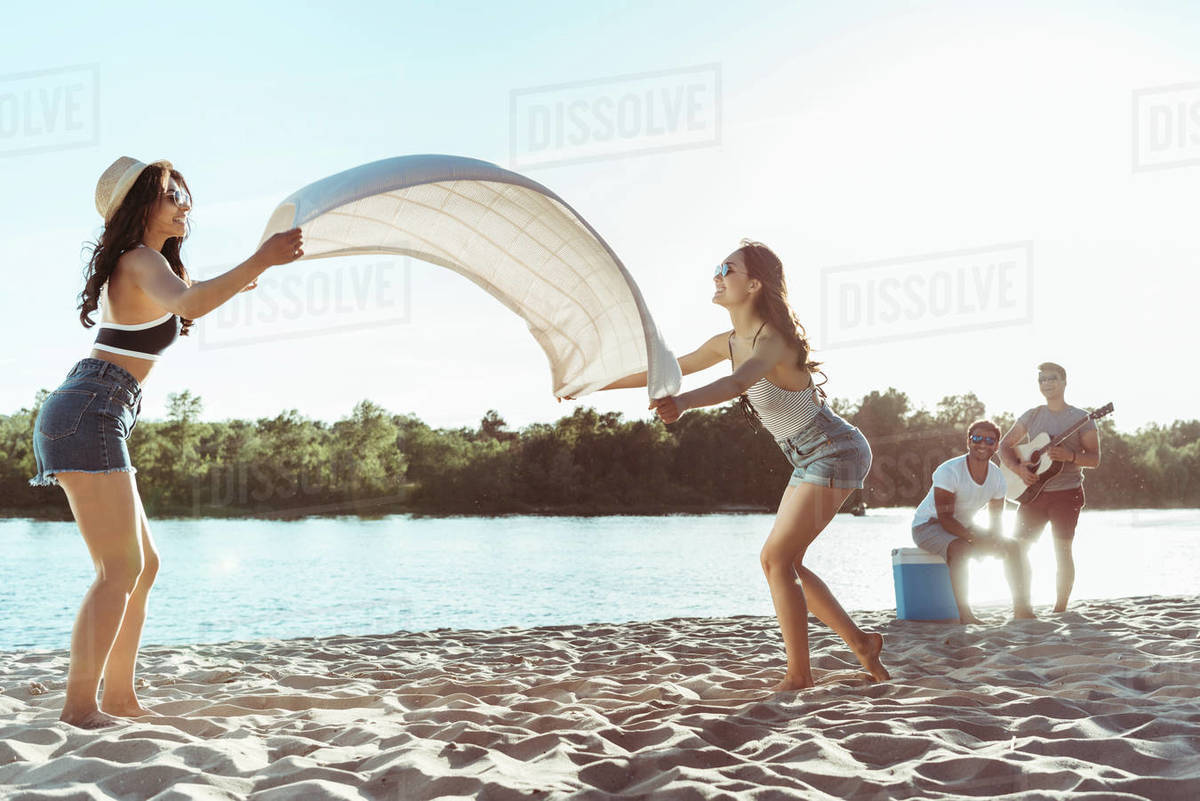 Attractive girls waving beach blanket on sandy riverside at daytime