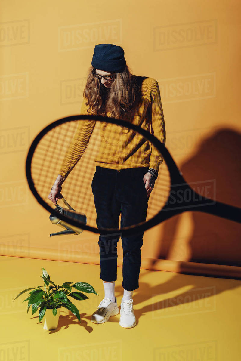 Stylish man watering plant, view through tennis racket on yellow