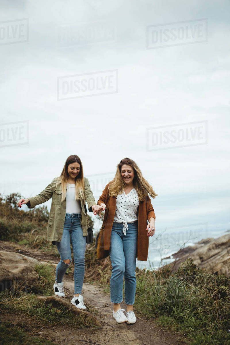 Cheerful young women holding hands and laughing while walking on narrow ...
