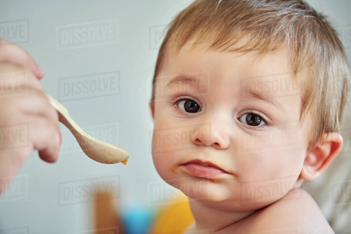 Closeup of adorable little baby eating puree from spoon held by crop