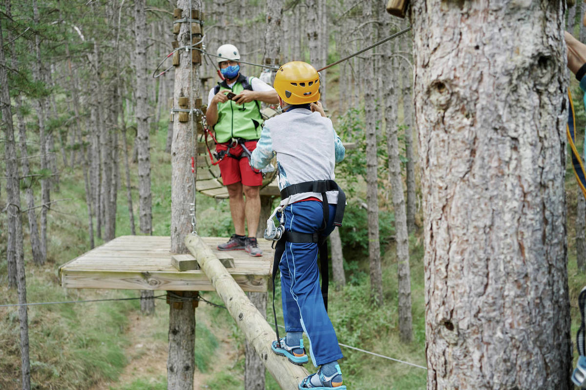 Unrecognizable boy in helmet using safety rope to walk on log towards man while climbing obstacle course in adventure park in forest Royalty-free stock photo