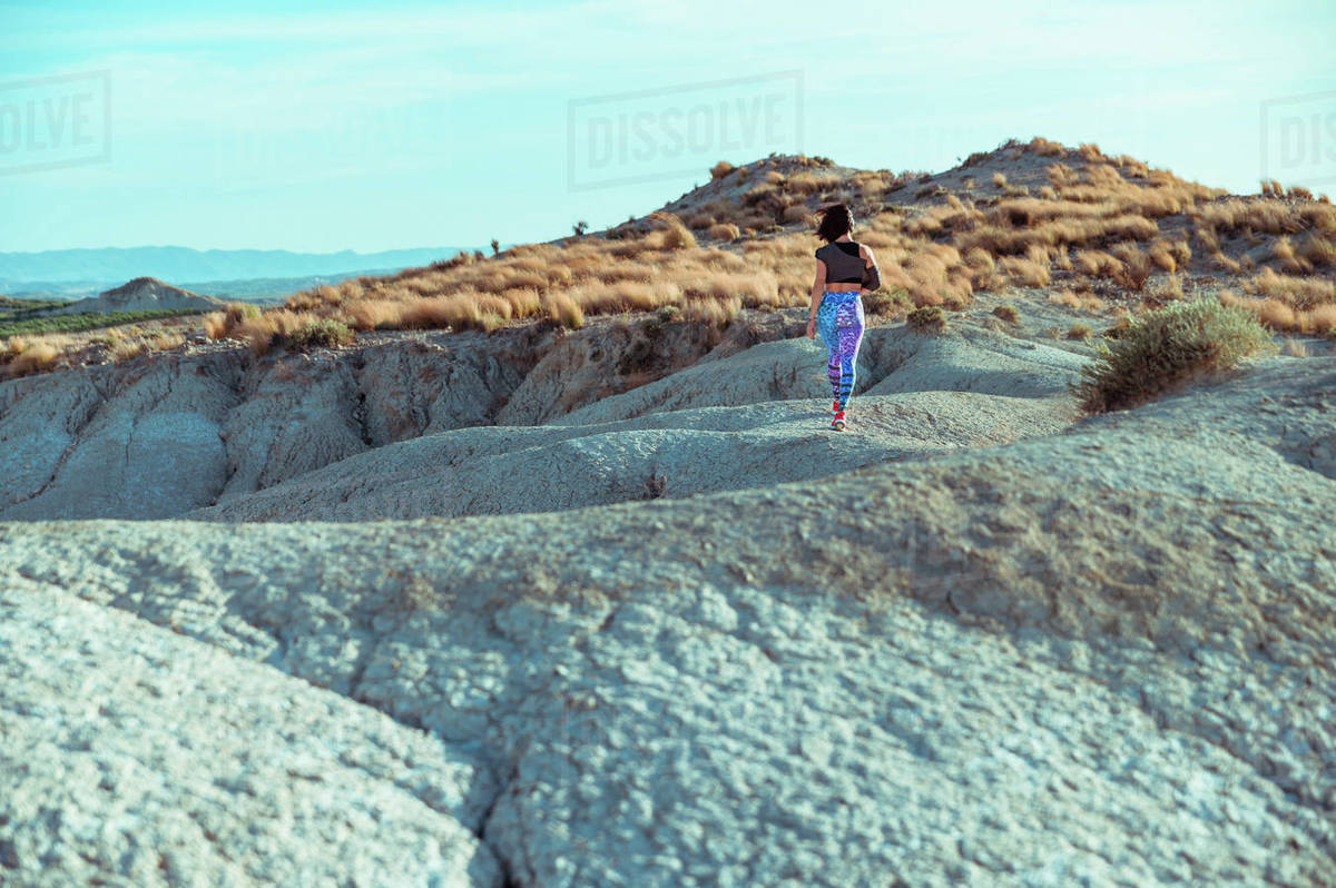 Back view of unrecognizable active female athlete in sportswear walking on rocky slope of rough desert hilly terrain under blue sky Royalty-free stock photo