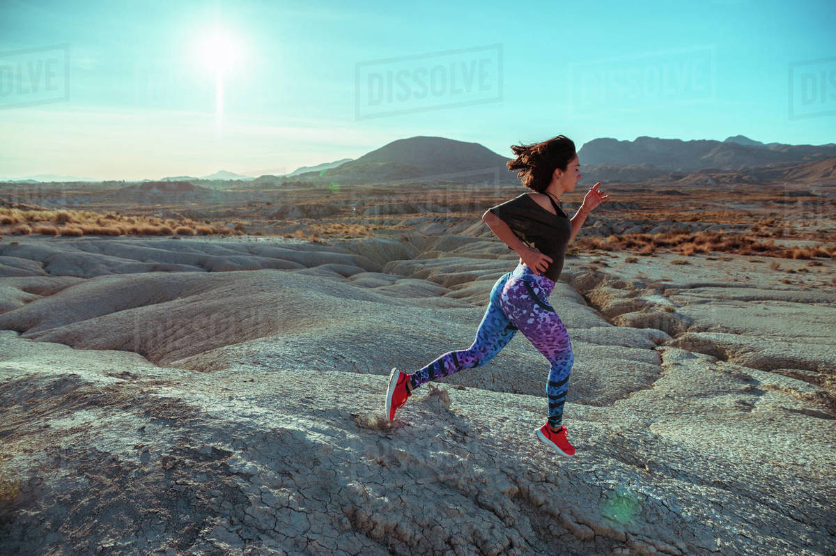 Full body side view of active female athlete in sportswear running and jumping over rocky slope of rough desert hilly terrain under blue sky Royalty-free stock photo