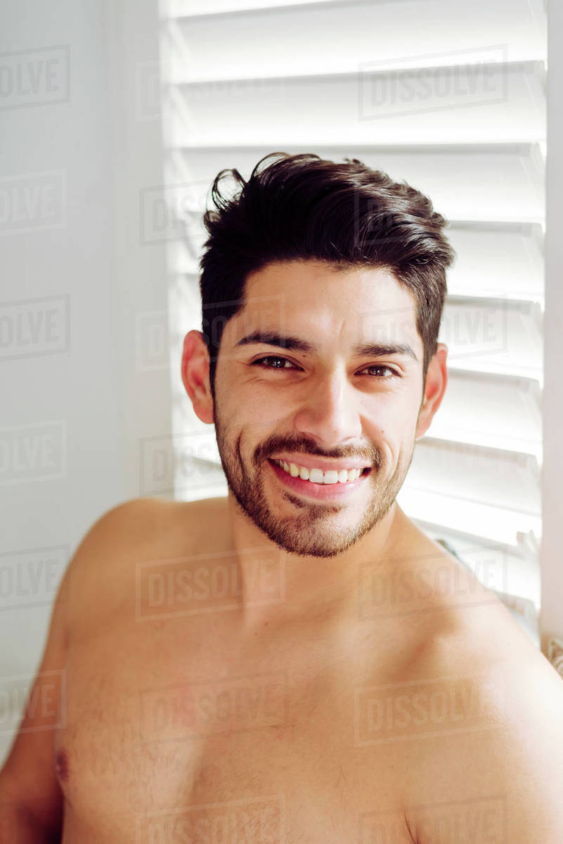 Young joyful naked handsome male with stylish hairstyle leaning on window at home and looking at