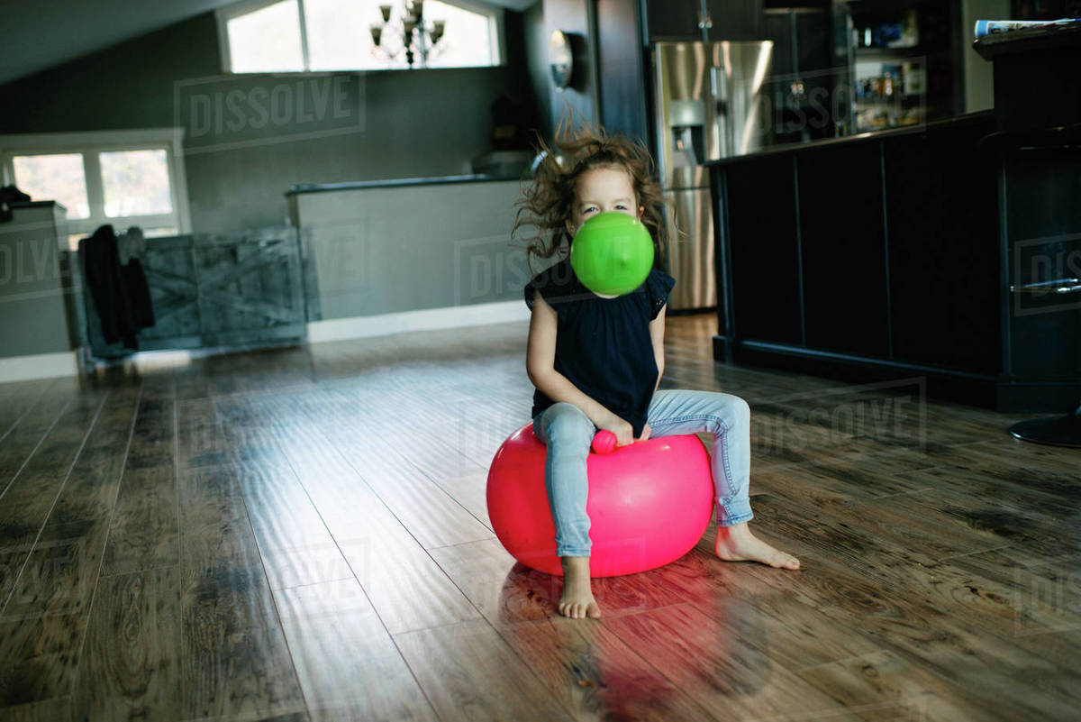 Little girl with balloon in mouth bouncing on gym ball at home Stock