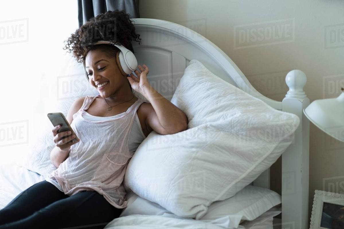 Woman relaxing on bed, wearing headphones, looking at smartphone