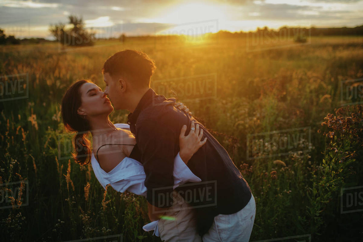 Young couple in love embraces and kisses on meadow at sunset. Backlit. Royalty-free stock photo