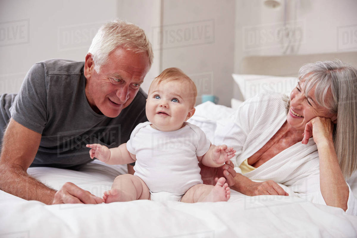 Grandparents Lying In Bed At Home Looking After Baby Grandson Stock