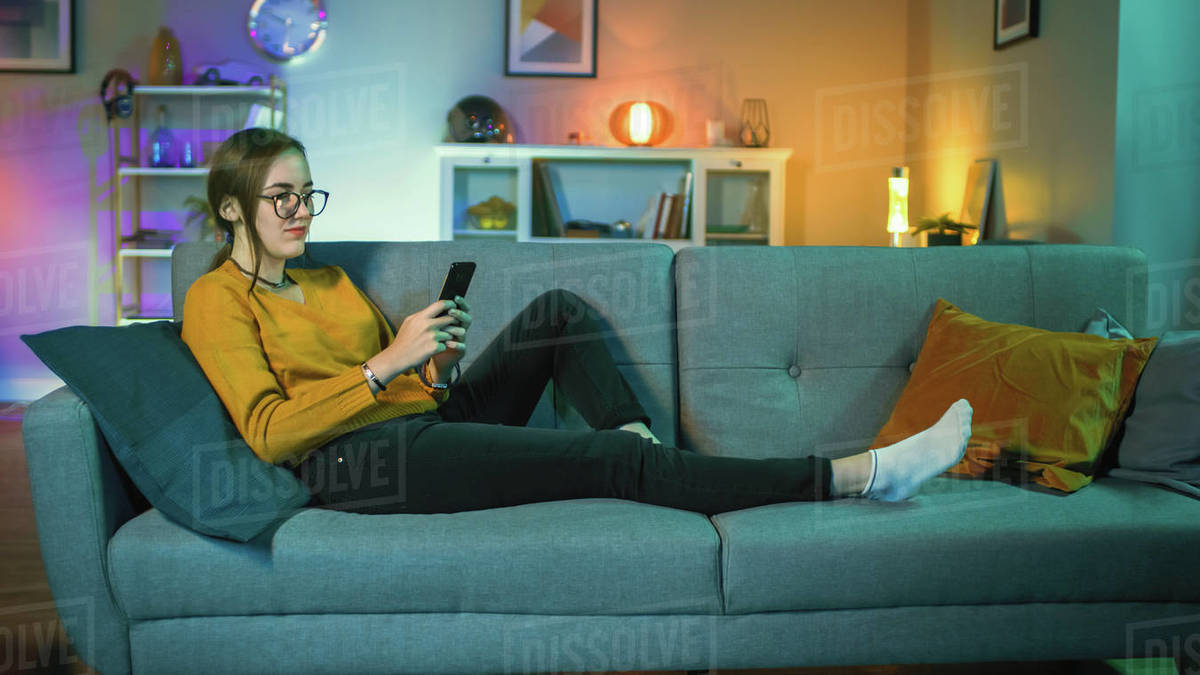 Beautiful excited young girl in glasses sitting on a couch and using her smartphone. She is smiling while tv plays in the background. Screen adds reflections to her face. Cozy room is lit with warm light. Royalty-free stock photo