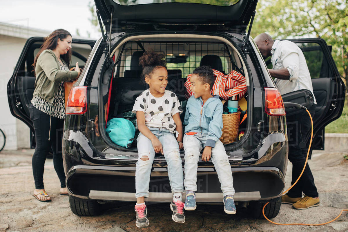 Full length of smiling siblings sitting on car trunk with parents standing in front yard Royalty-free stock photo