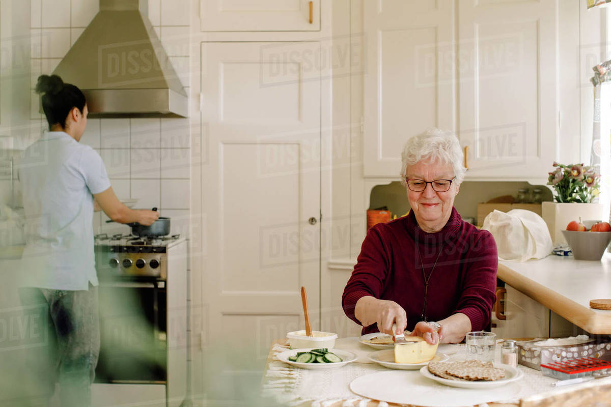 Smiling retired elderly woman eating breakfast while female caregiver