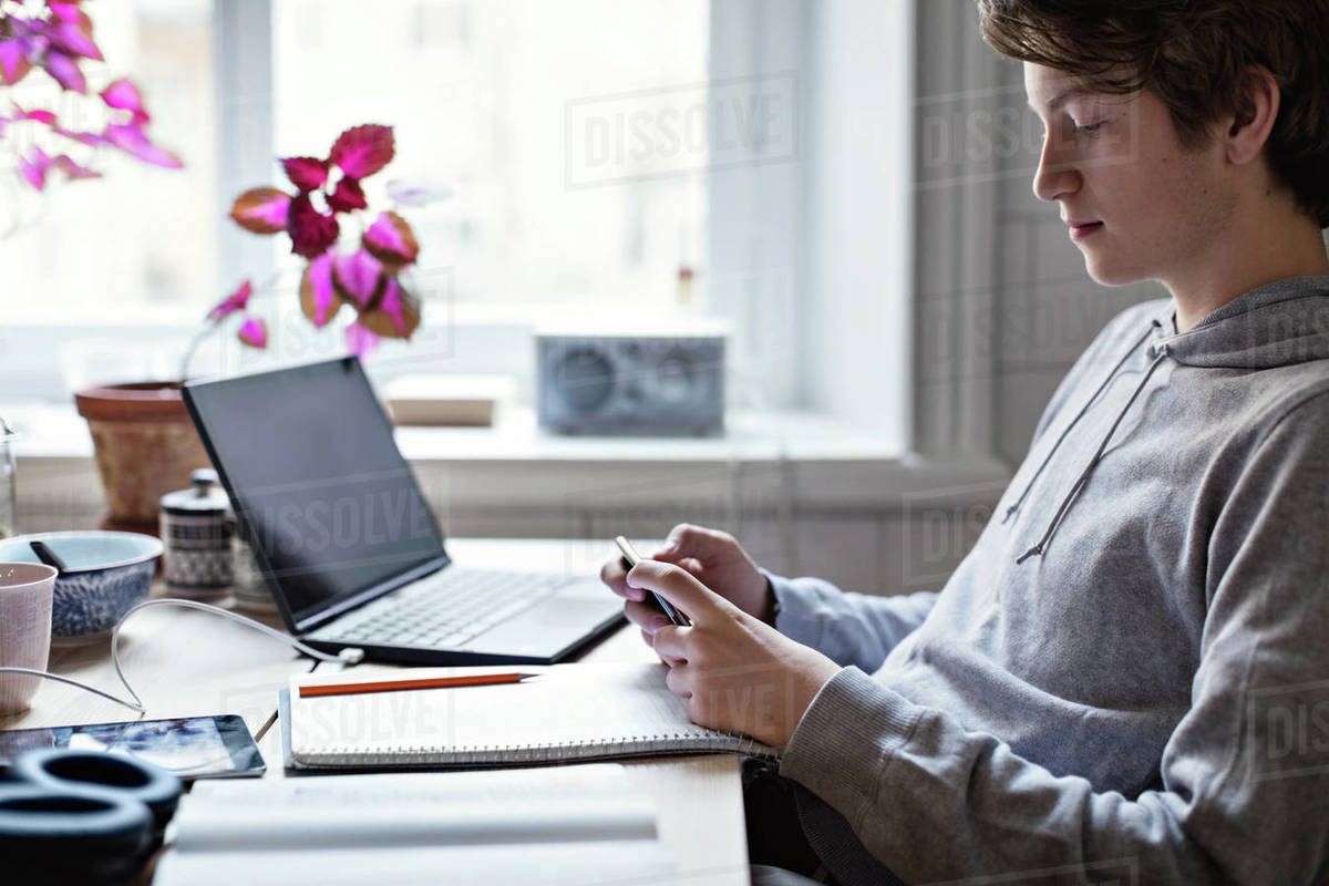 Side view of teenage boy using mobile phone while sitting with books and laptop on table at home Royalty-free stock photo