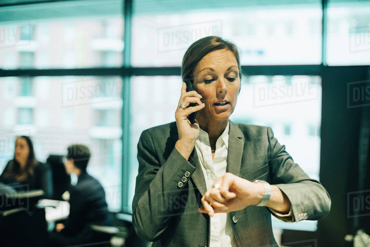 Busy businesswoman talking on mobile phone while checking time on watch