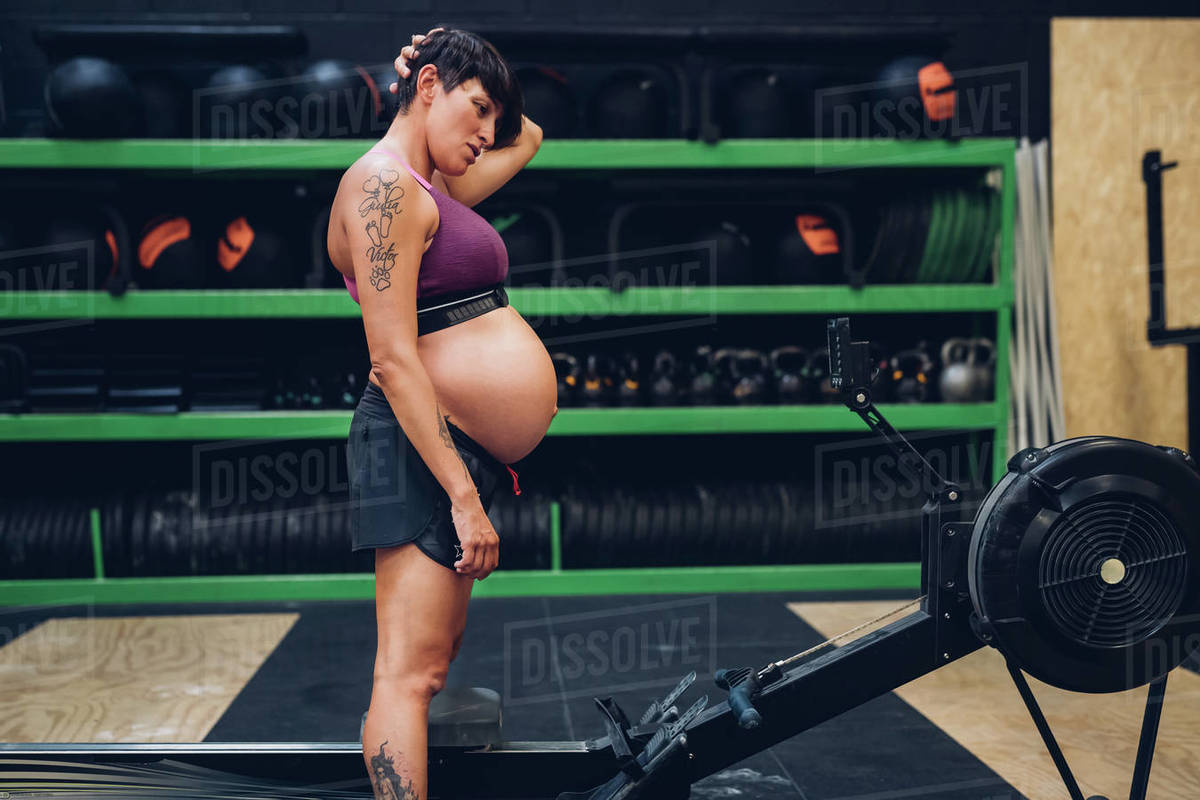 Pregnant woman looking at rowing machine in gym Stock Photo Dissolve