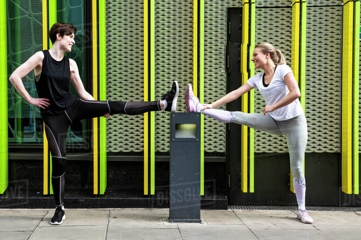 Two young female runners stretching legs on city sidewalk, full length Royalty-free stock photo