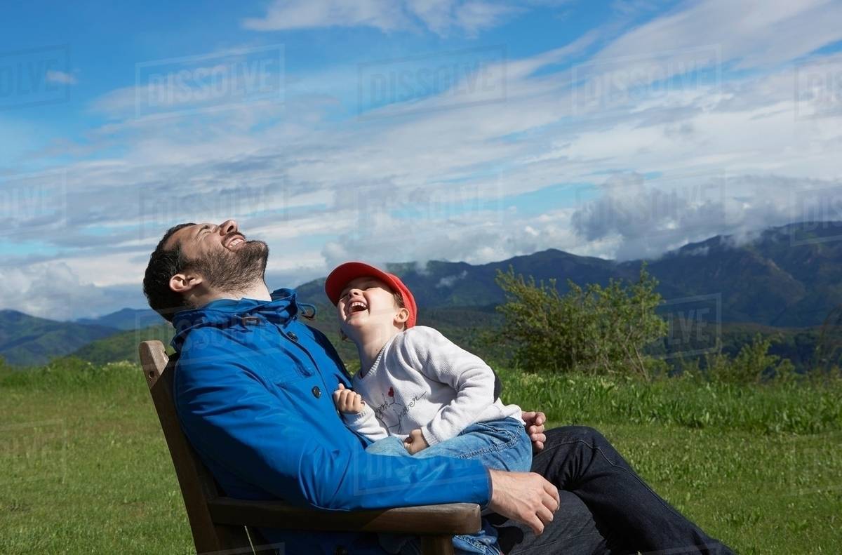 Father and daughter sitting on chair outdoors laughing Stock Photo