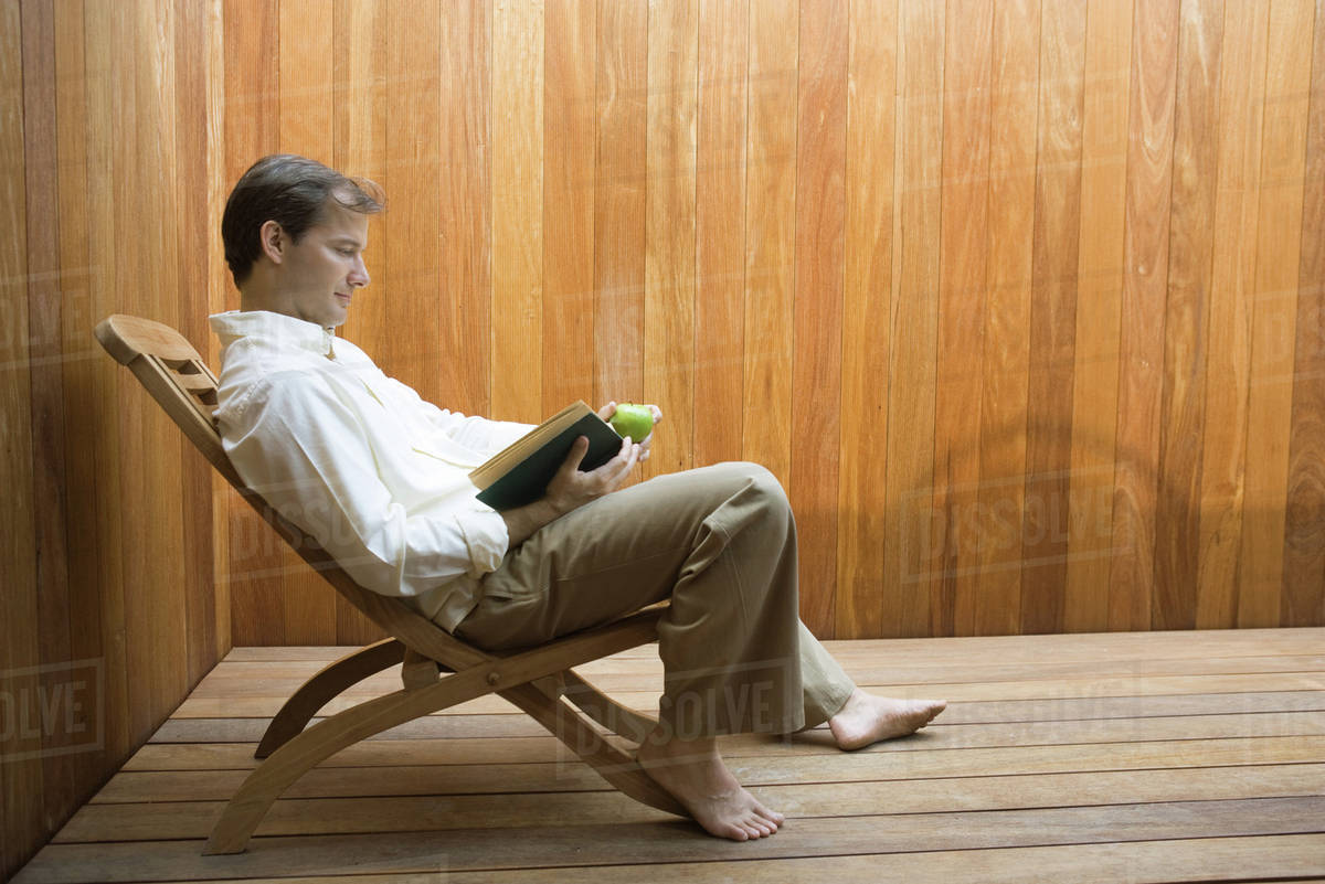 Man sitting in lounge chair reading book, apple in hand, side view