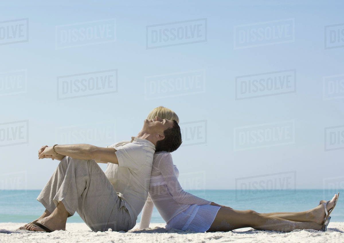 Couple sitting back to back on beach Stock Photo Dissolve