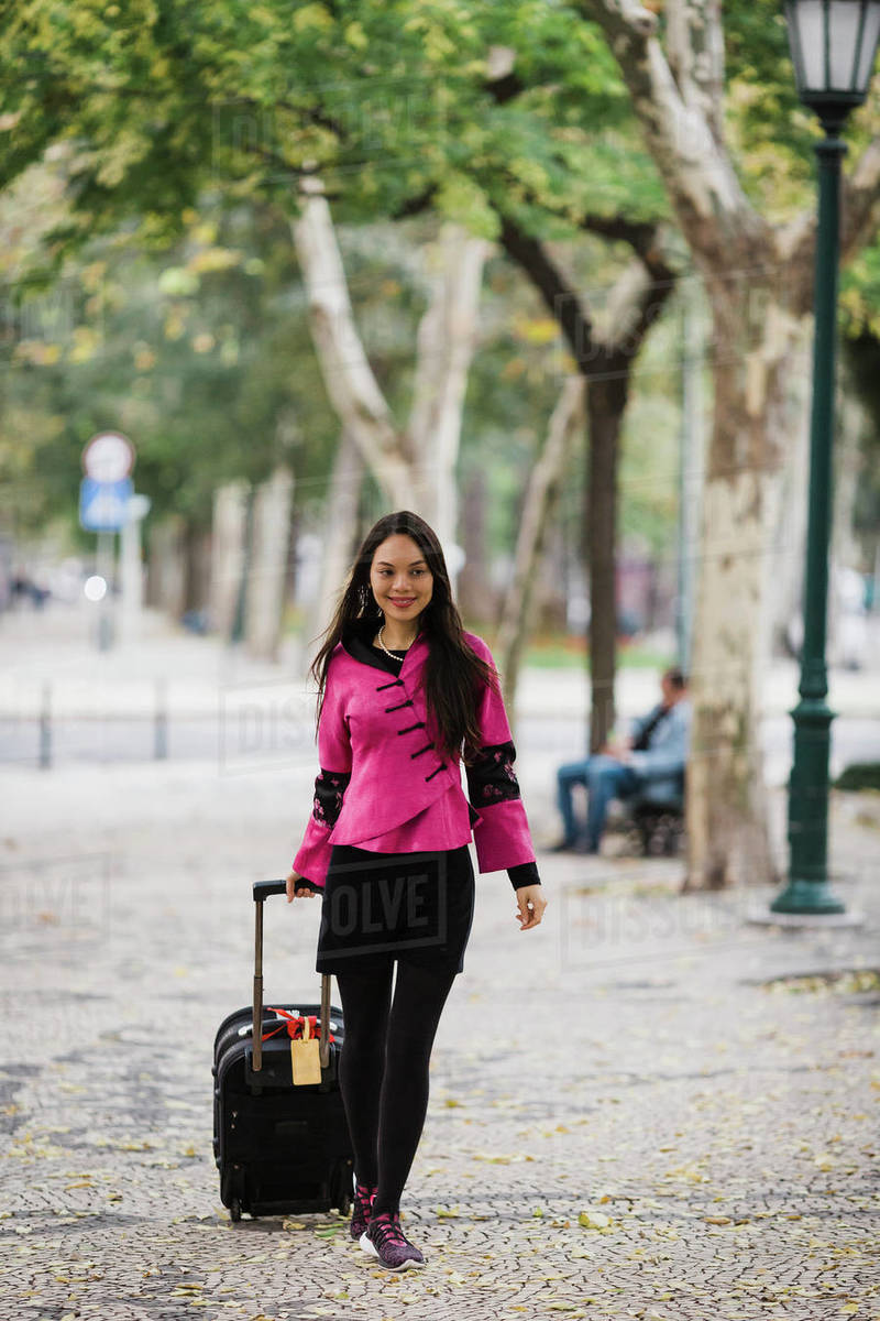 Smiling female tourist pulling suitcase along treelined sidewalk Royalty-free stock photo